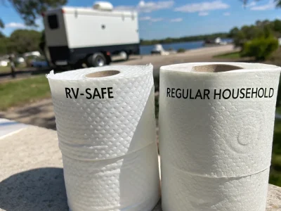 Two toilet paper rolls labeled 'RV-Safe' and 'Regular Household' on a ledge with an RV and lake in the background.