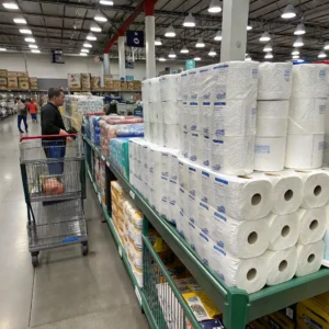 A person shopping in a large wholesale store with shelves stacked with paper products and a shopping cart in the aisle.