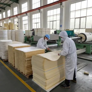Two workers in protective clothing handle sheets of paper in an industrial paper manufacturing plant, surrounded by stacks of paper and machinery.